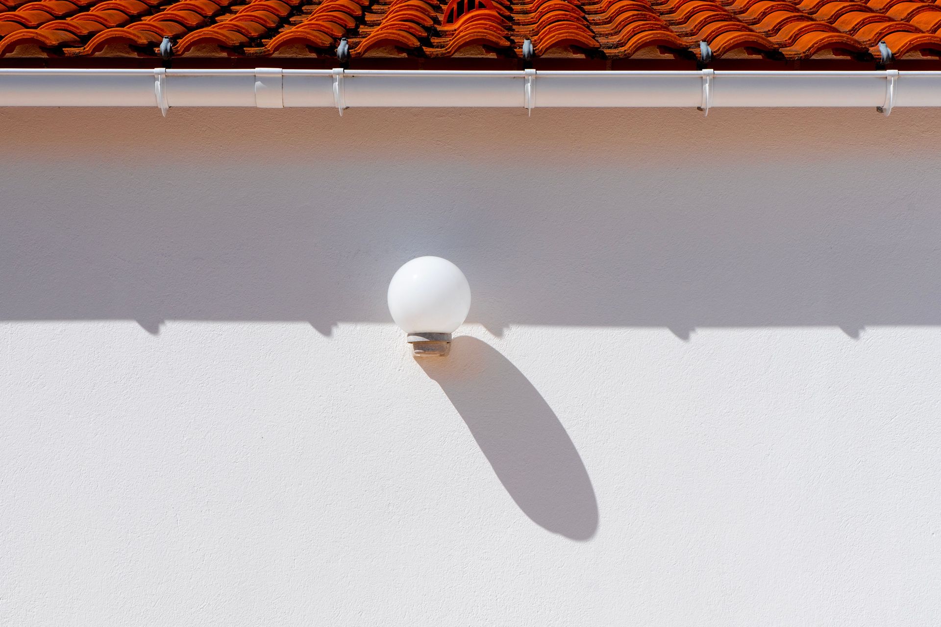 A minimalist outdoor setting showcasing a wall lamp casting a shadow beneath a red tiled roof.